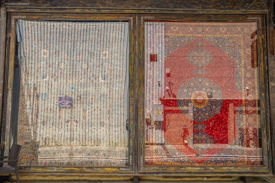 A side-by-side comparison of two oriental rugs displayed behind large glass windows in a commercial space. The left rug is visibly aged and stained, with faded colors and worn fibers, indicating a nee