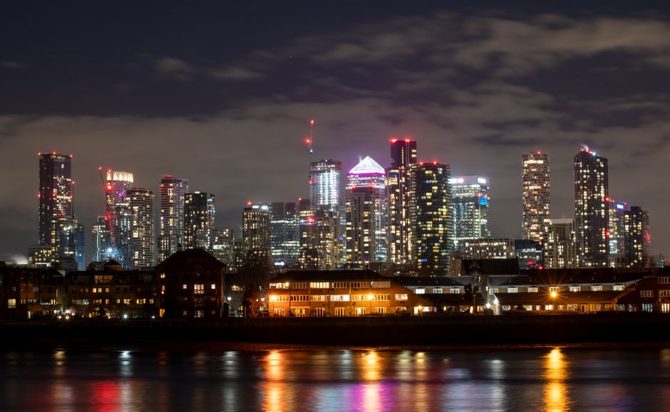 Nighttime view of the Canary Wharf skyline reflected on the calm river surface, showcasing illuminated modern office buildings with glass and steel exteriors, some featuring distinctive lighting accen